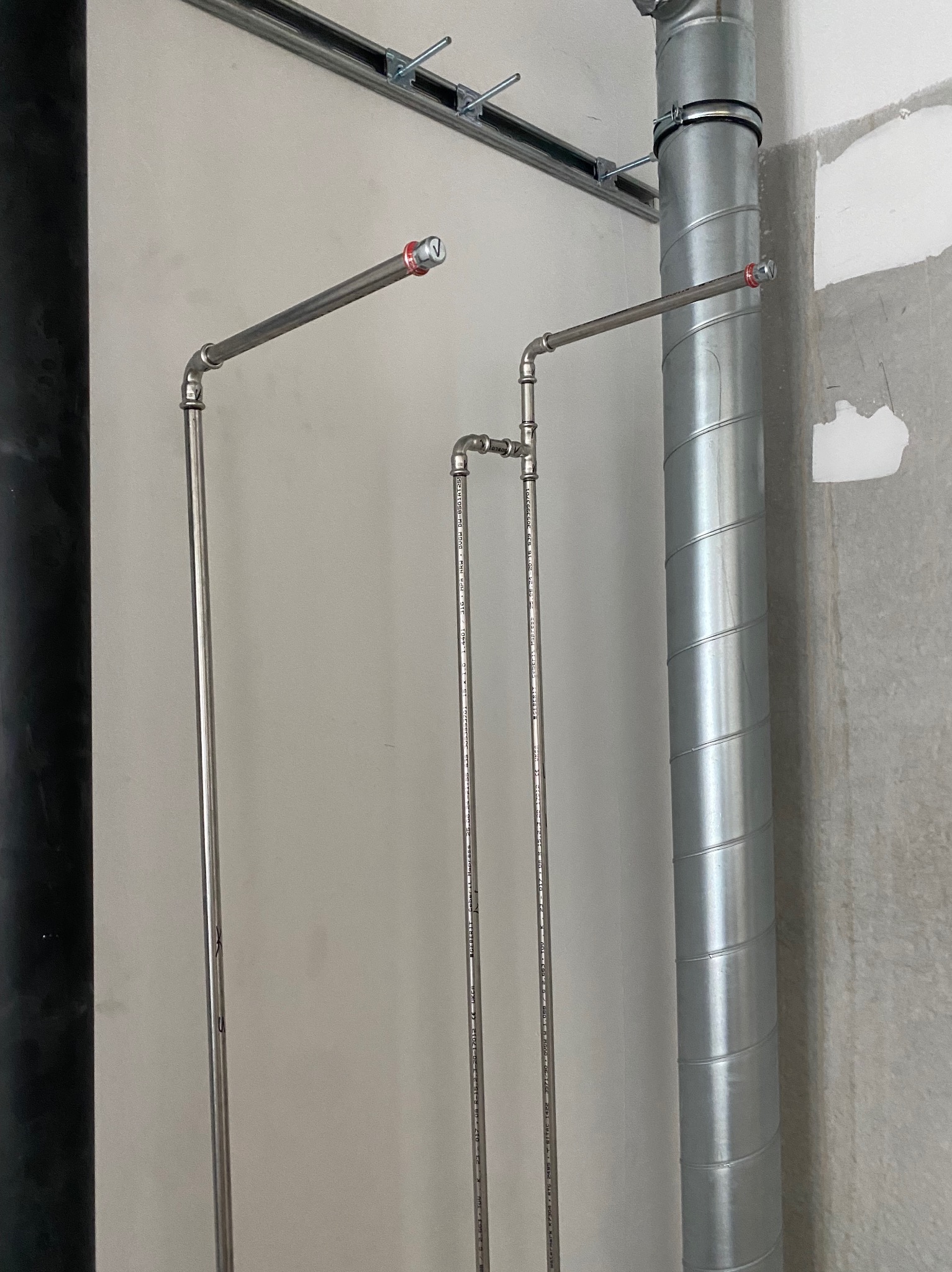 Szczegół instalacji: rury ze stali nierdzewnej, pionowe i poziome, z elementami złącznymi, obok spiralnej rury wentylacyjnej na tle surowej ściany.