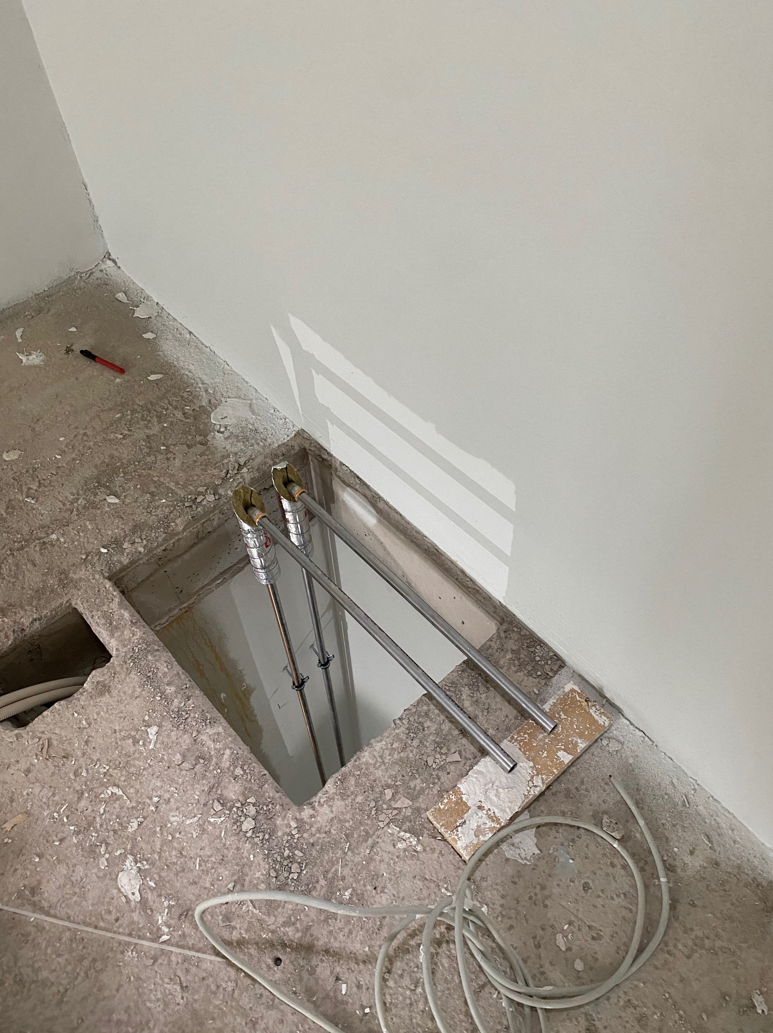 Fragment instalacji hydraulicznej z rurami w otworze w podłodze, zabezpieczonymi izolacją. Ściana biała, widoczne kable i fragment podłogi w trakcie prac.