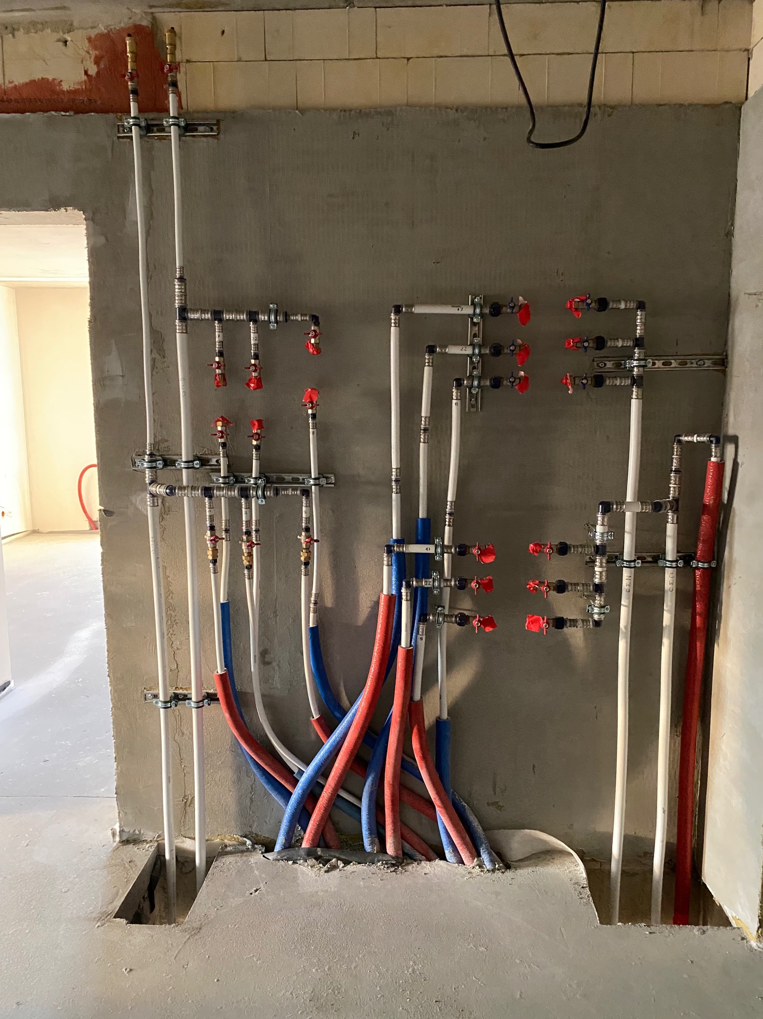 Węzeł instalacji wodnej w surowym stanie: białe rury pionowe, czerwone i niebieskie poziome, liczne zawory z czerwonymi pokrętłami, szare ściany i podłoga w trakcie remontu.