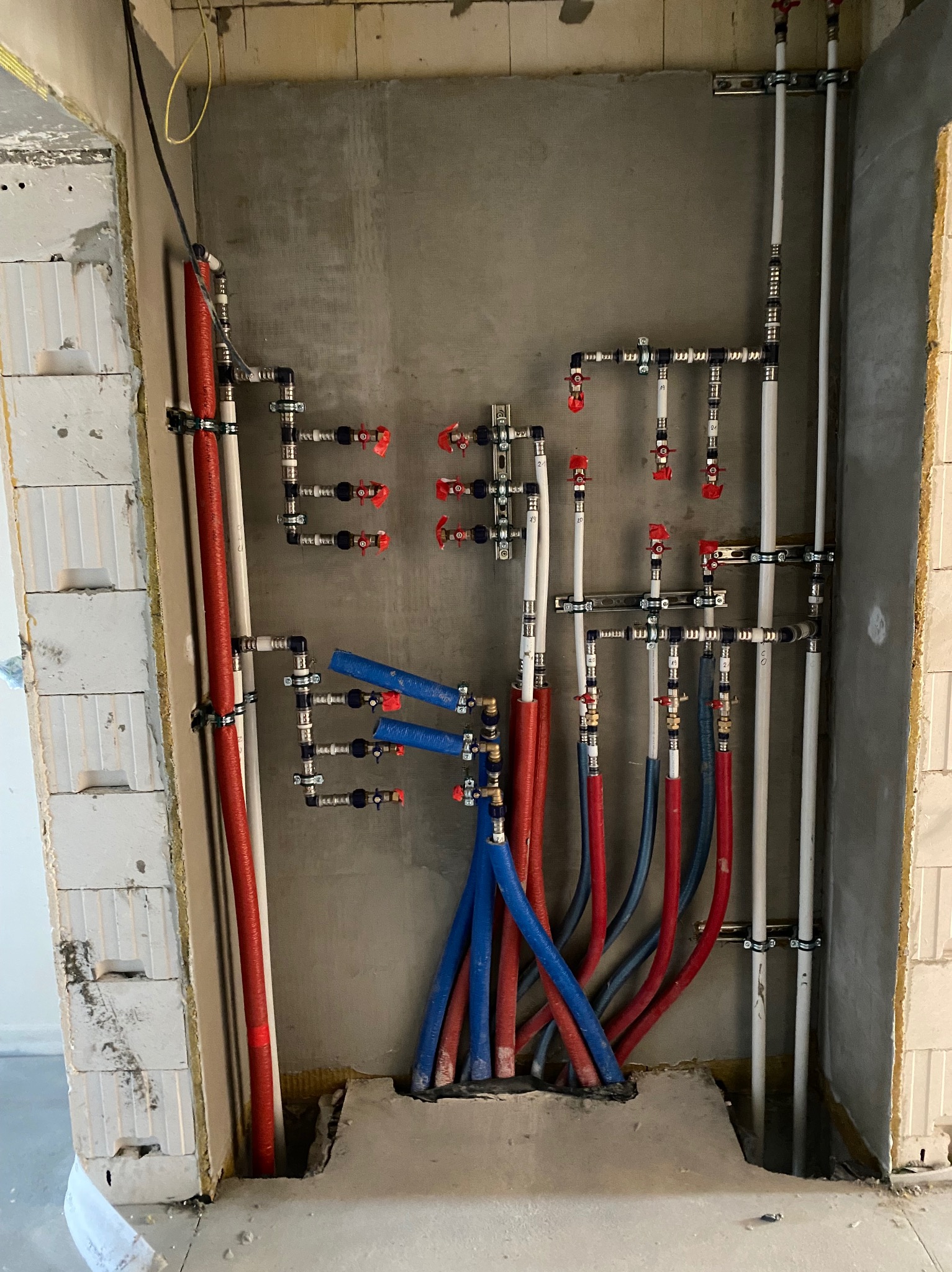 Instalacja wodno-kanalizacyjna w surowym stanie: czerwone i niebieskie rury PEX, białe rury PCV i zawory zamontowane na szarej ścianie z betonu komórkowego.