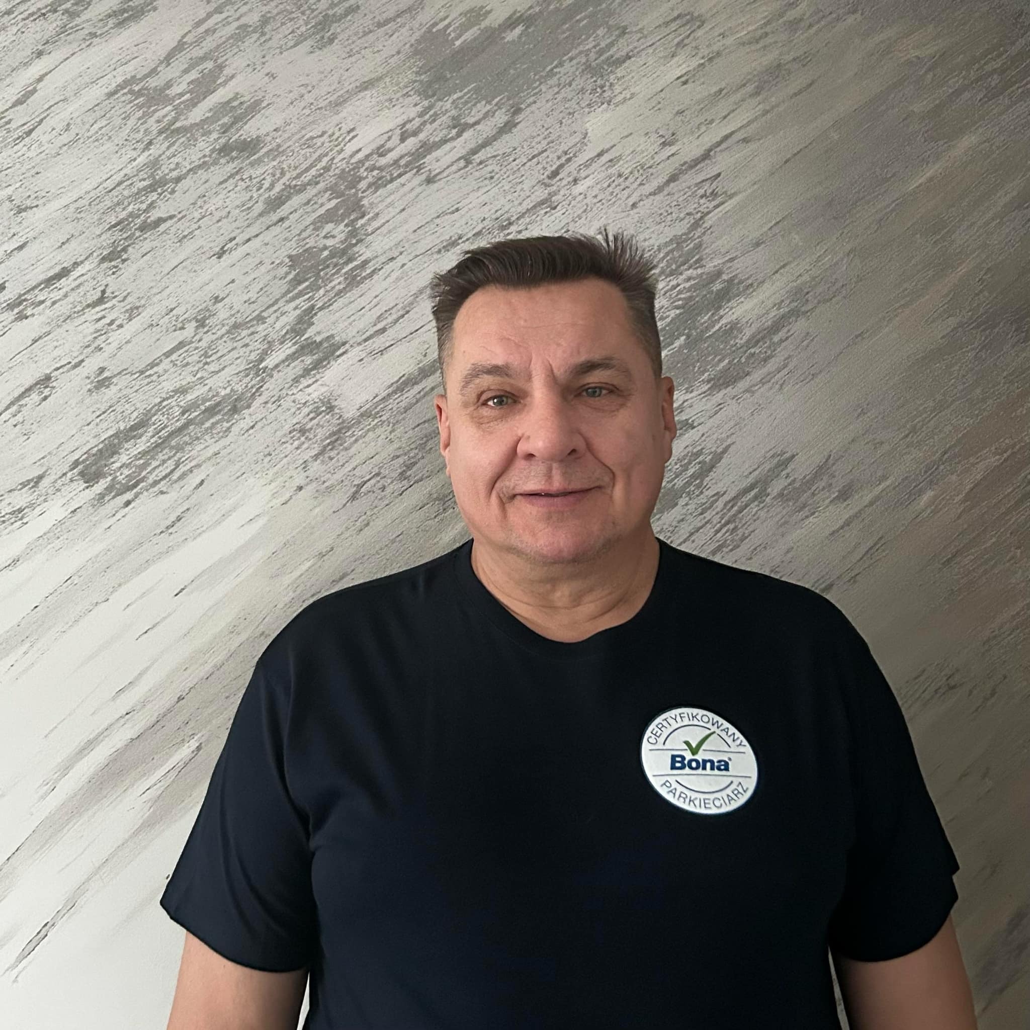 Portret certyfikowanego parkieciarza Bona na tle ściany z efektem dekoracyjnym. Mężczyzna w ciemnej koszulce z logo Bona.