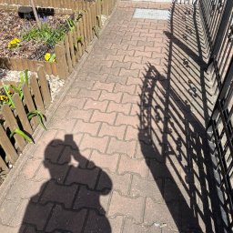 Mobilna Myjna SV - Czerwona kostka brukowa przed domem w Stargardzie, z cieniem ogrodzenia i osoby robiącej zdjęcie. Widoczne schody wejściowe i fragment rabaty z kwiatami.