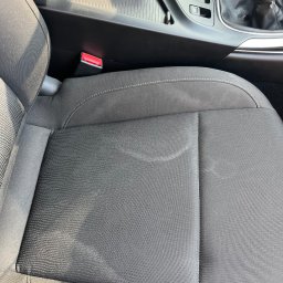 Mobilna Myjna SV - Szare, materiałowe siedzenie samochodowe z widocznymi plamami przed praniem tapicerki. Widoczny fragment konsoli środkowej i pas bezpieczeństwa.