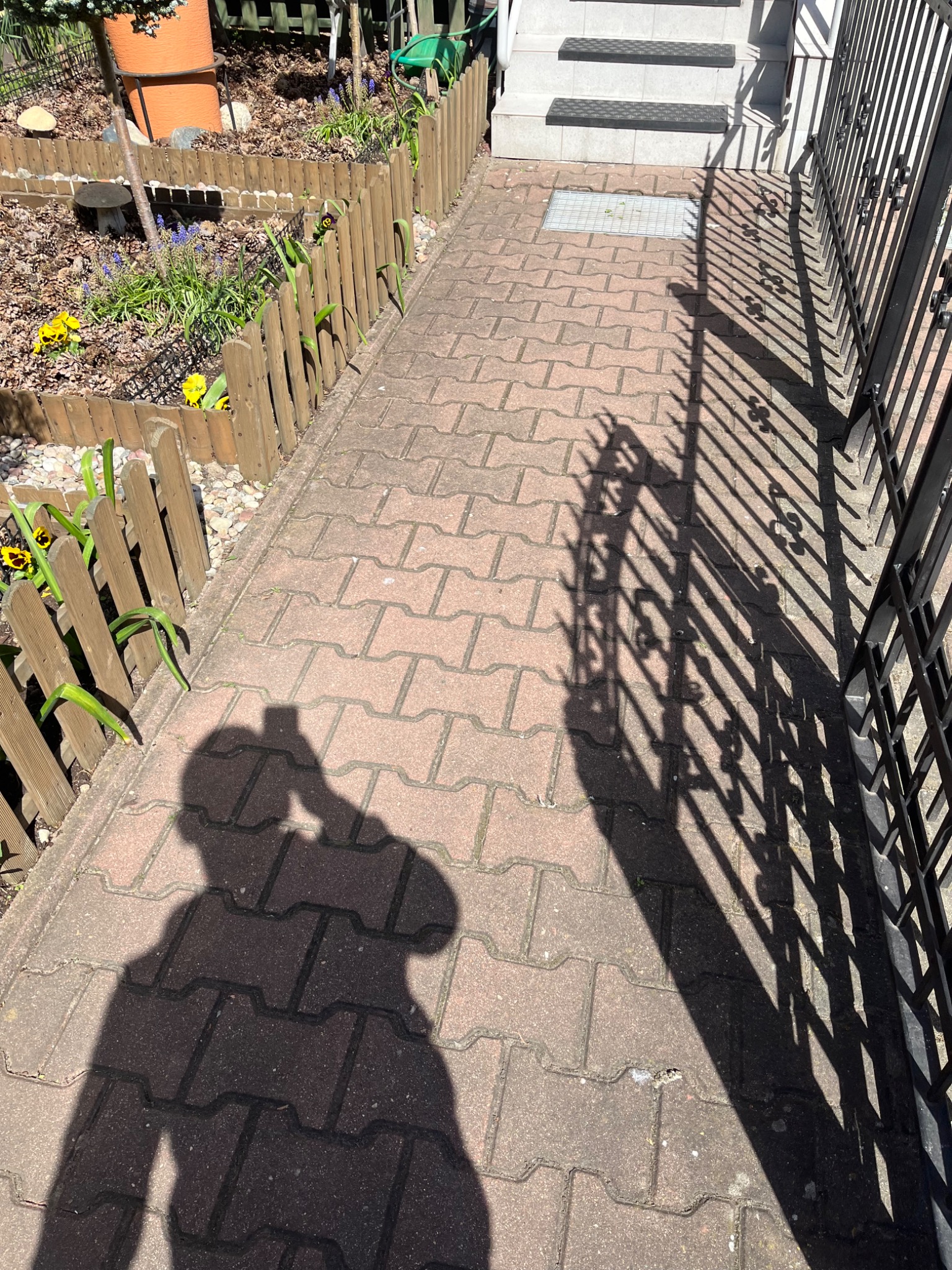 Czerwona kostka brukowa przed domem w Stargardzie, z cieniem ogrodzenia i osoby robiącej zdjęcie. Widoczne schody wejściowe i fragment rabaty z kwiatami.