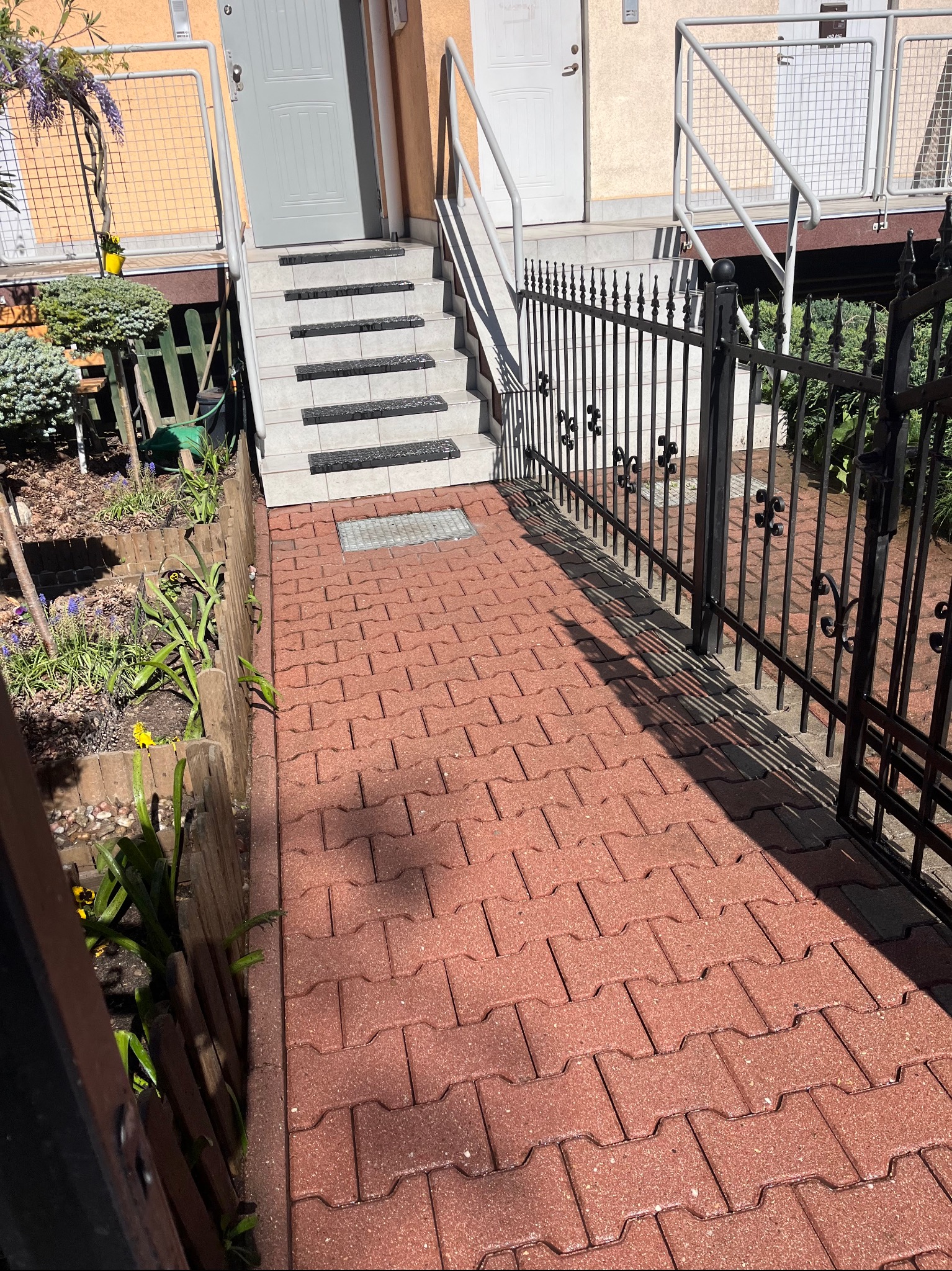 Czerwona kostka brukowa prowadzi do schodów z czarno-białymi akcentami. Metalowe ogrodzenie i bujna zieleń otaczają ścieżkę.