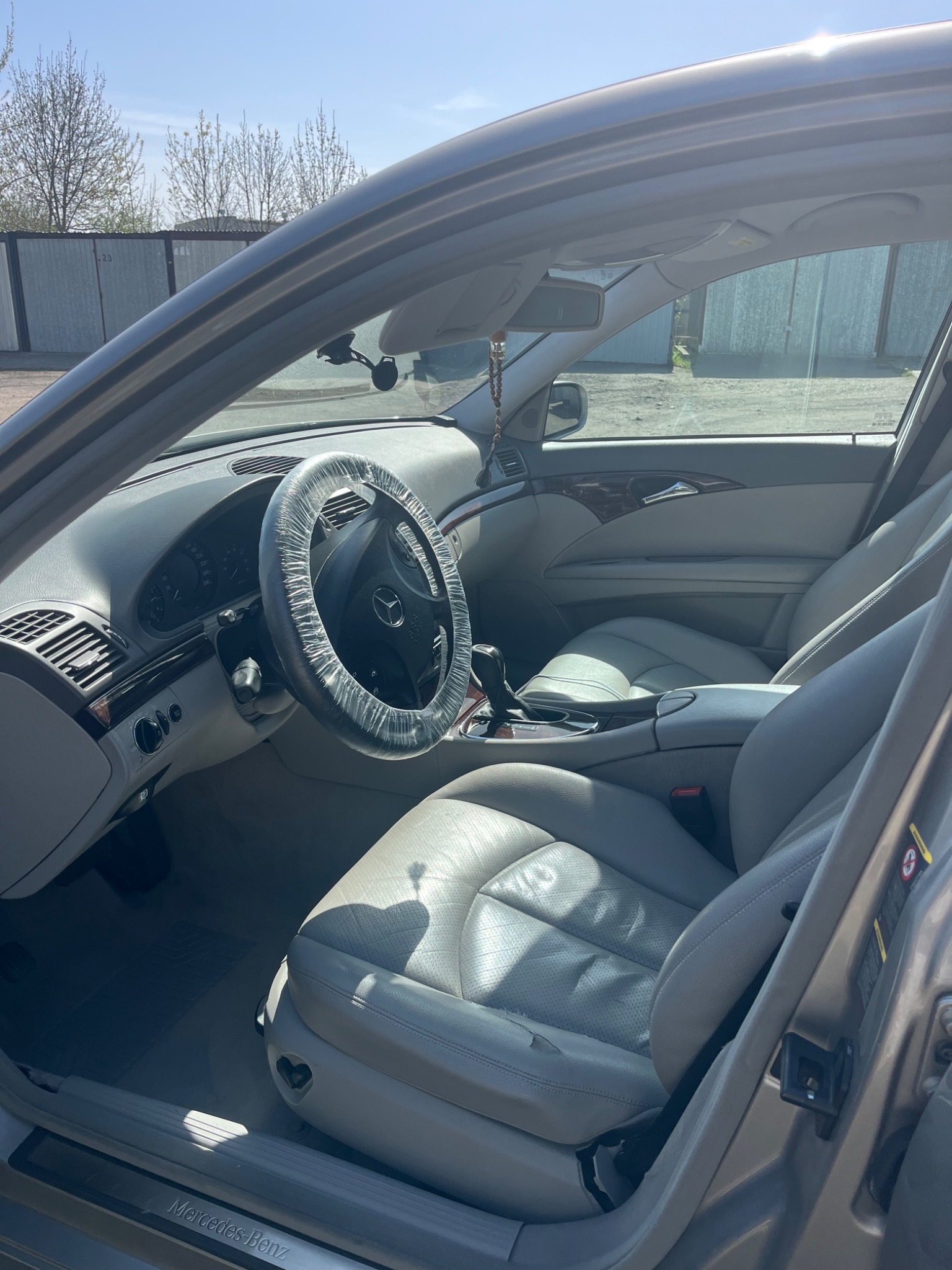 Wnętrze srebrnego Mercedesa E-Klasy; skórzana tapicerka w odcieniach szarości, kierownica owinięta folią ochronną, widok z zewnątrz przez otwarte drzwi.