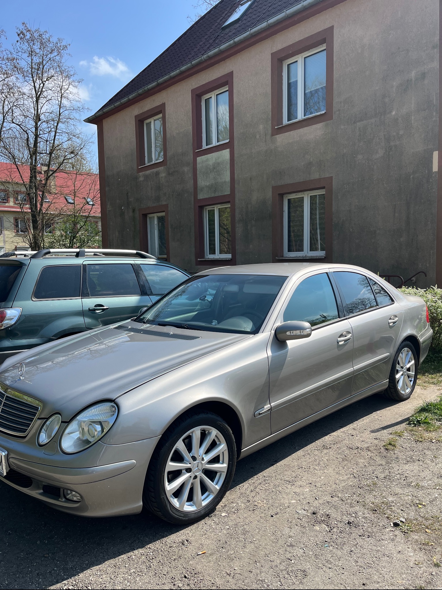 Jasnoszary Mercedes-Benz E-Klasa W211 na żwirowym podjeździe przed budynkiem z szarą elewacją i białymi oknami. Drugi samochód w tle.