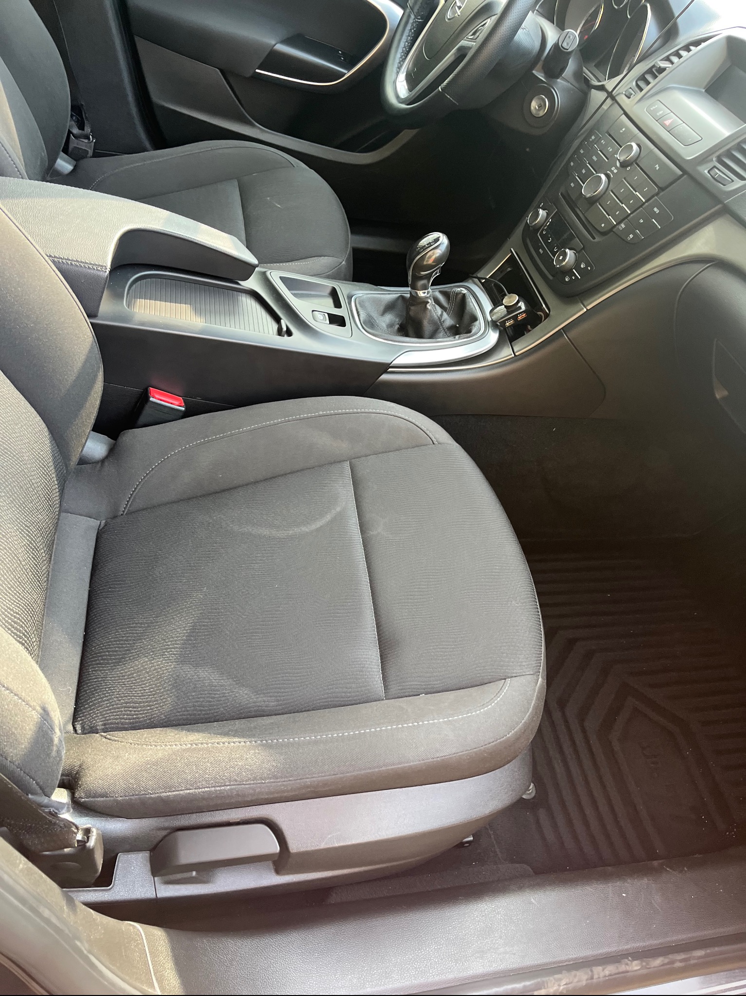 Wnętrze samochodu z widocznym siedzeniem kierowcy z plamą, konsola środkowa z dźwignią zmiany biegów i panel sterowania. Dywaniki samochodowe z wzorem.