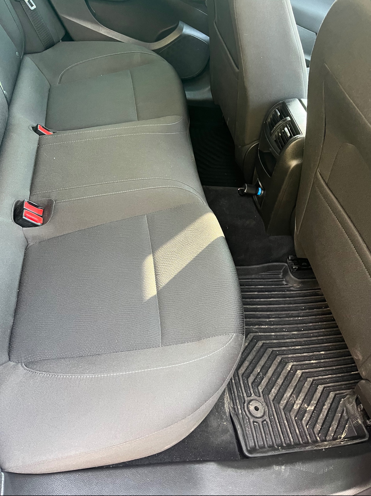 Wnętrze samochodu: tylna kanapa z szarej tapicerki, czarne dywaniki gumowe z widocznymi zabrudzeniami, gniazdo ładowania. Widok z perspektywy pasażera.