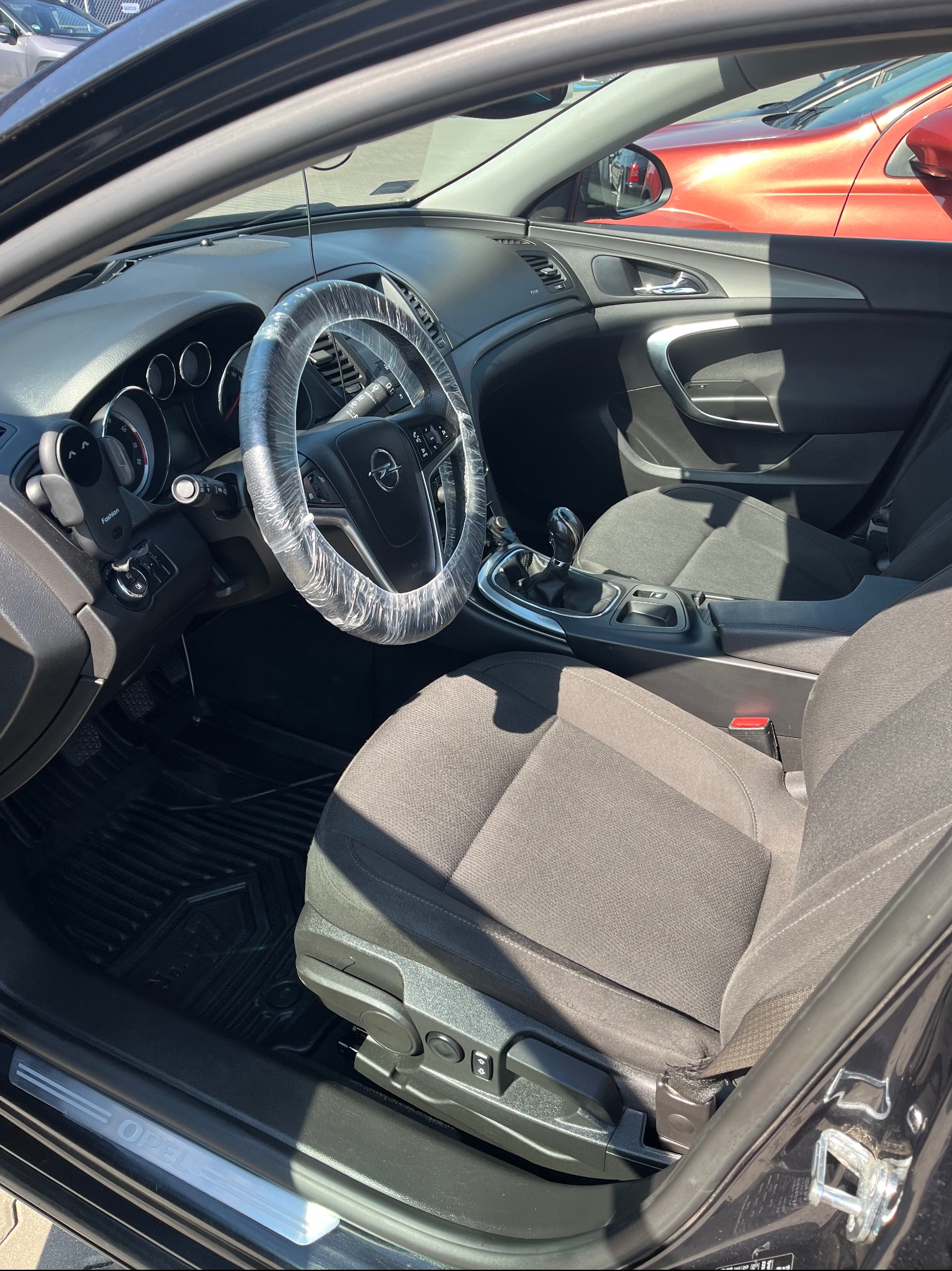 Wnętrze czarnego Opla Insignia: szare fotele, kierownica owinięta folią ochronną, deska rozdzielcza i elementy wyposażenia. Widok z zewnątrz auta.