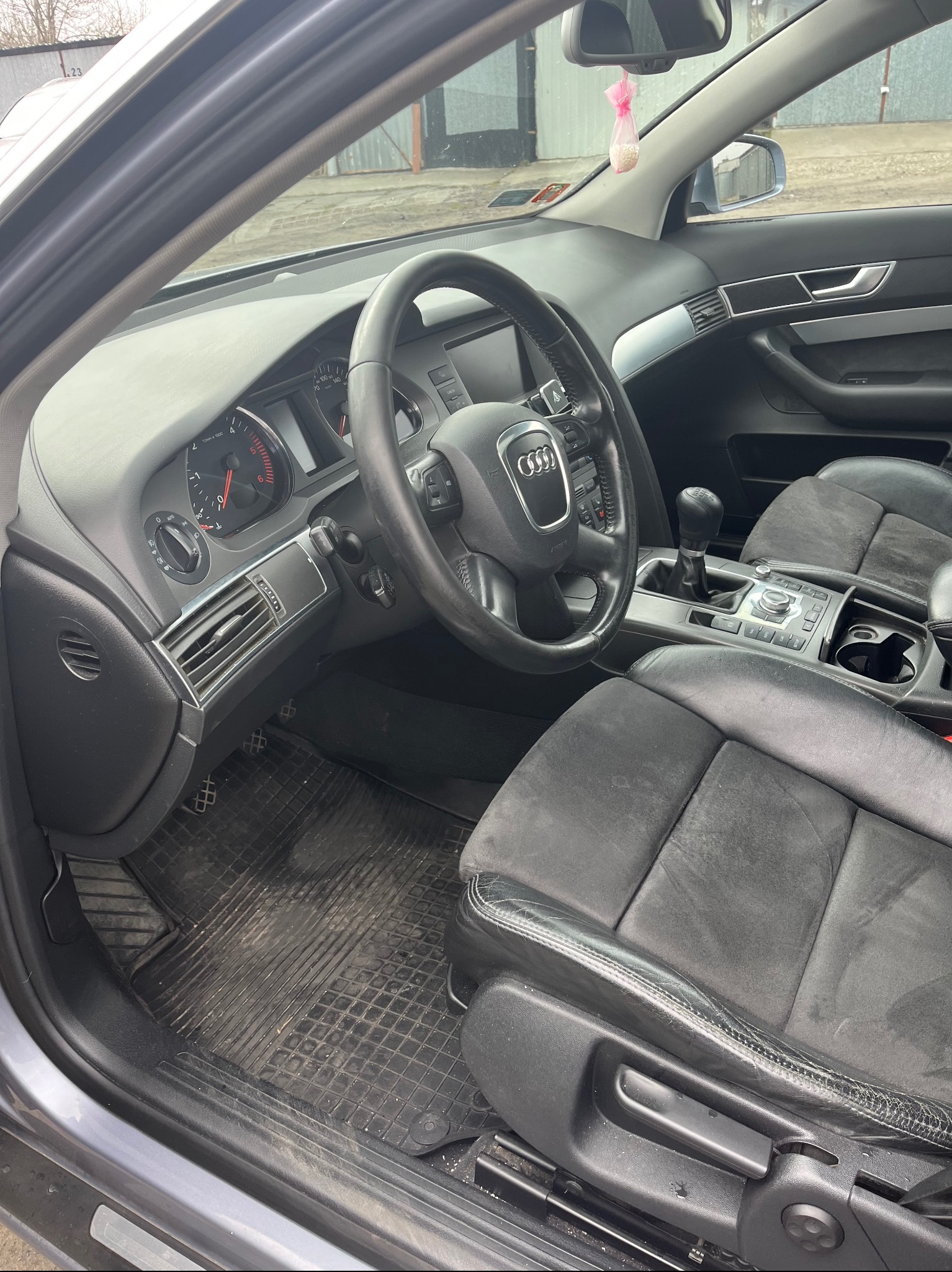 Wnętrze samochodu Audi A6: czarne, skórzane fotele, widoczna deska rozdzielcza z zegarami, kierownica z logo Audi i brudna gumowa wycieraczka pod nogami kierowcy.