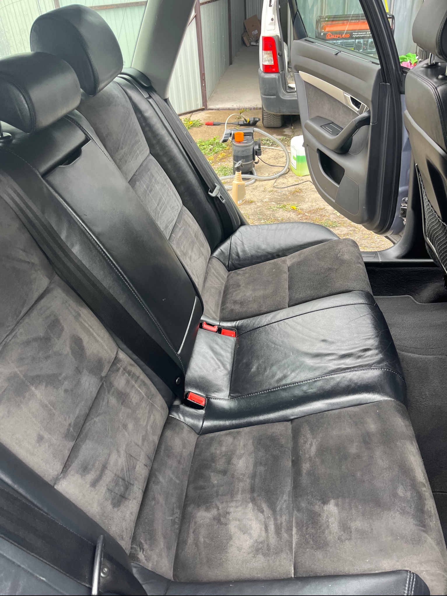 Wnętrze samochodu z tylną kanapą po czyszczeniu; widoczne odświeżone, czyste obicia skórzane i zamszowe oraz urządzenie do prania tapicerki w tle.