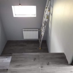 RemontUp Mariusz Sękowski - Jasne płytki na schodach i szara ściana z oknem dachowym. Biały grzejnik i drabina oparta o ścianę. Ukończony remont klatki schodowej z wykończeniem poddasza.