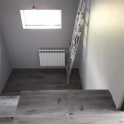 Jasne płytki na schodach i szara ściana z oknem dachowym. Biały grzejnik i drabina oparta o ścianę. Ukończony remont klatki schodowej z wykończeniem poddasza.