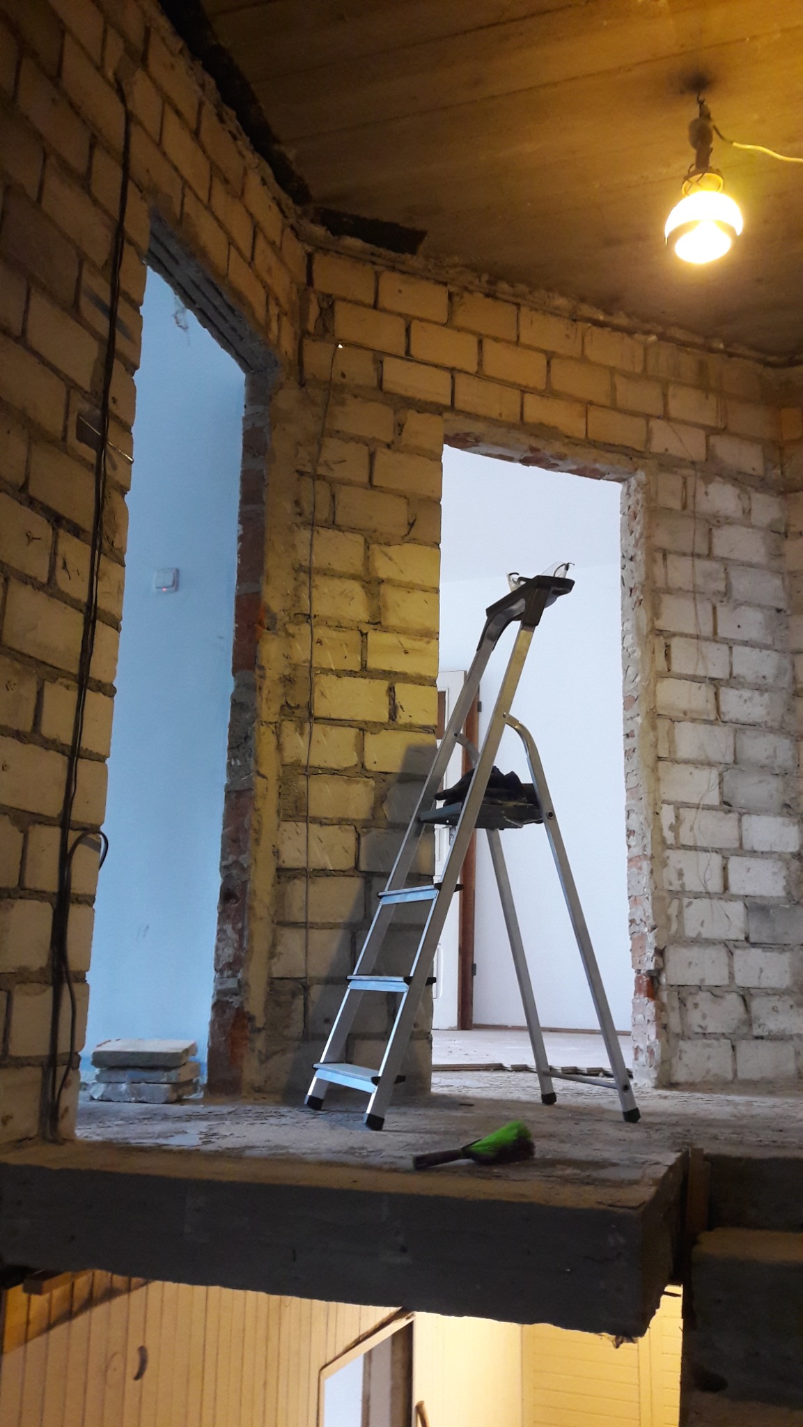 Surowy stan wnętrza z ceglanymi ścianami i aluminiową drabiną. Widoczne kable, surowy betonowy strop i prowizoryczne oświetlenie. Etap prac remontowych.