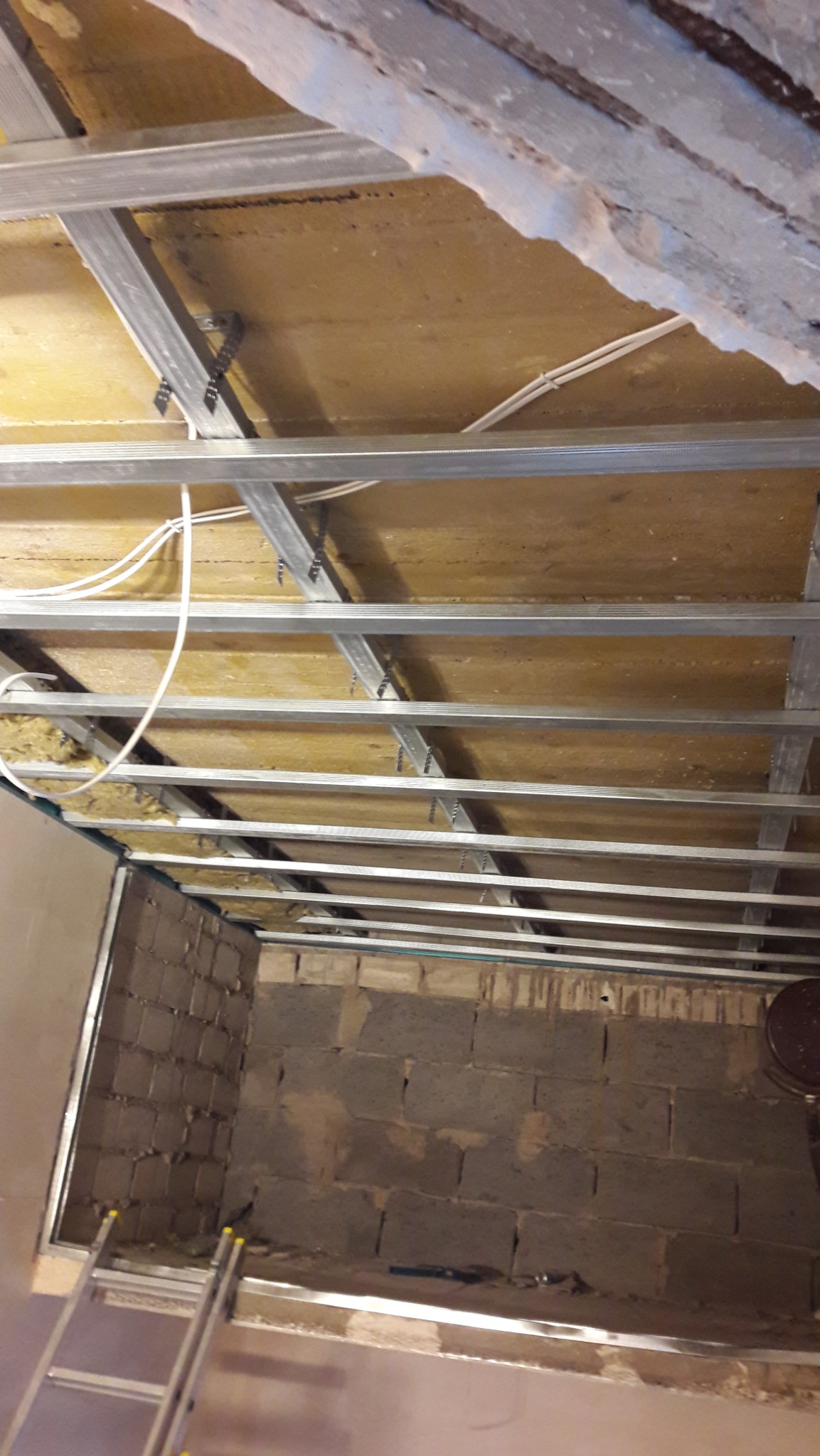 Montaż sufitu podwieszanego z widoczną izolacją i metalową konstrukcją. Ściany z szarej cegły, drabina w rogu. Przewody elektryczne pod sufitem.