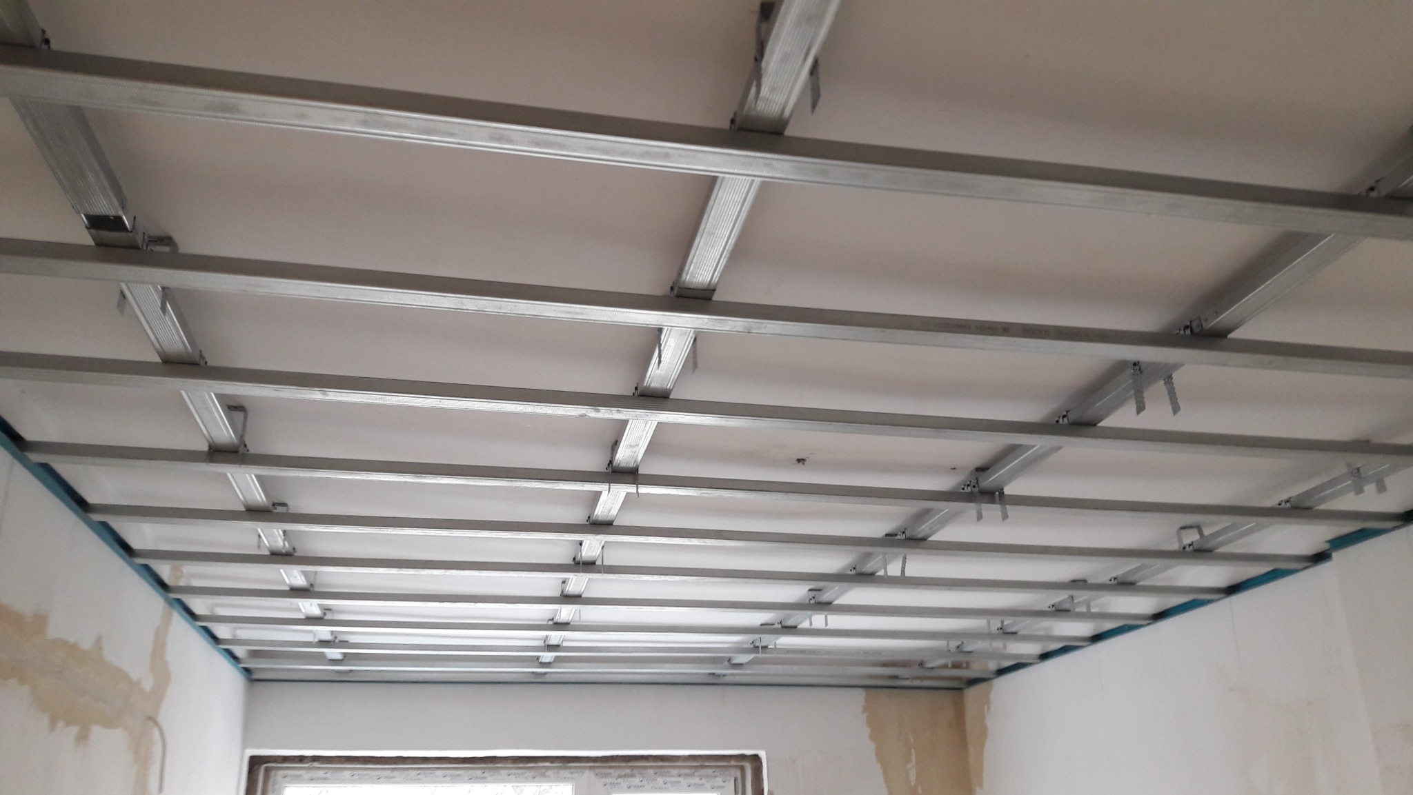Sufit podwieszany w trakcie montażu. Widoczne metalowe profile i płyty gipsowo-kartonowe. Ściany przygotowane do dalszych prac wykończeniowych.