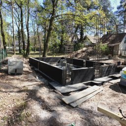 7Trust Igor Wełnowski - Fundamenty z bloczków betonowych i zbrojeniami, otoczone drzewami. W tle widoczny dom jednorodzinny. Etap budowy w naturalnym otoczeniu.