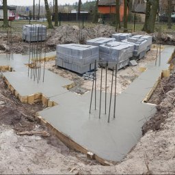 7Trust Igor Wełnowski - Zalane fundamenty domu z wystającymi prętami zbrojeniowymi i bloczkami betonowymi w folii. Widok z góry na wykopane fundamenty w ziemi.