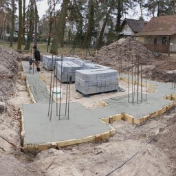 7Trust Igor Wełnowski - Wylane fundamenty pod budowę domu, widoczne szalunki, zbrojenie i bloczki betonowe na paletach. W tle domy i drzewa.