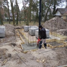 7Trust Igor Wełnowski - Fundamenty domu w Lubiczu: wylewanie betonu z pompy do szalunków zbrojeniowych. Na placu budowy widoczne bloczki i ziemia.