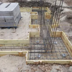 Szczegółowy widok na szalunek fundamentów zbrojonych stalą, przygotowany do wylania betonu. Na pierwszym planie paleta bloczków fundamentowych.