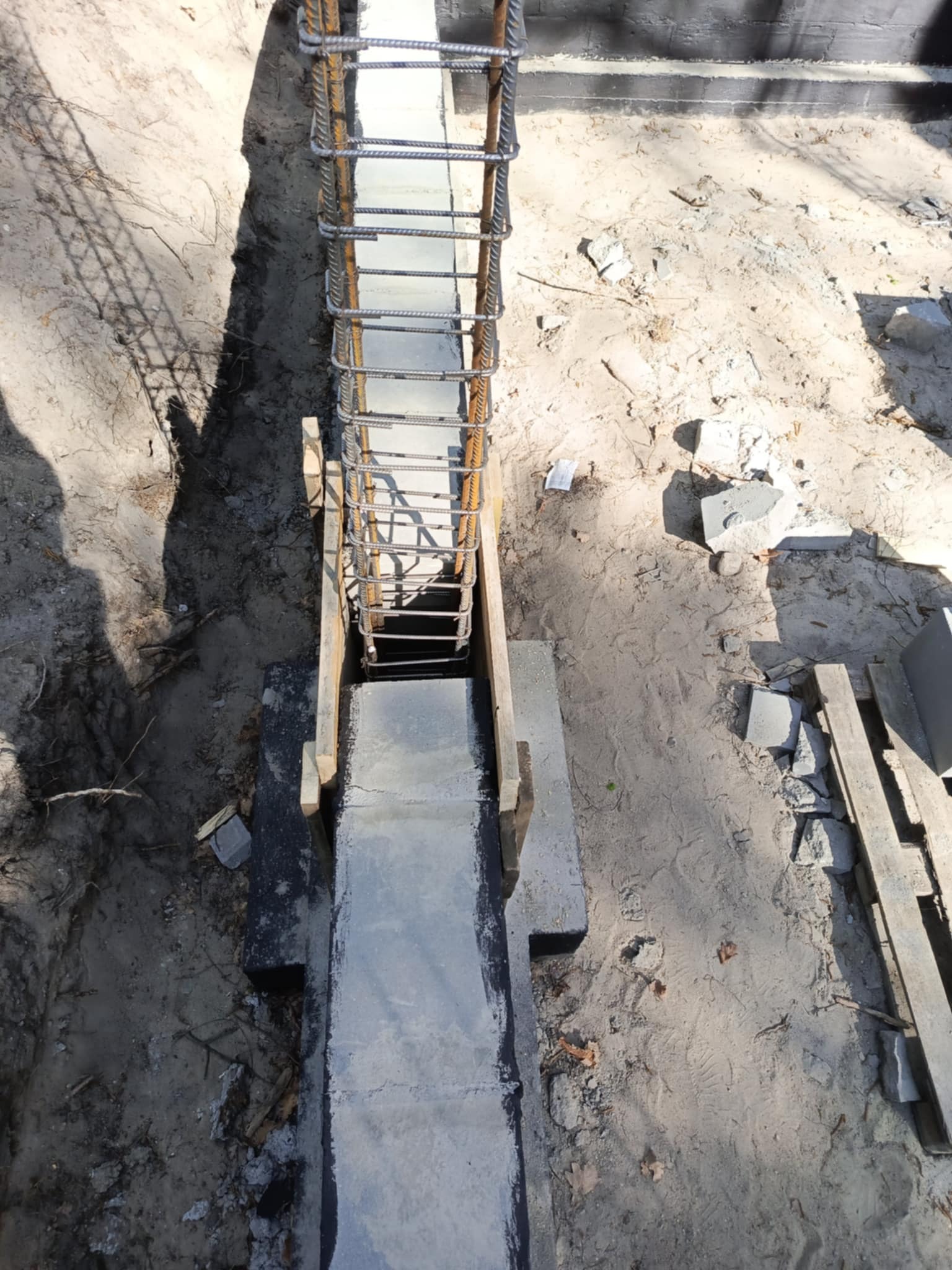 Zbrojenie fundamentu w wykopie, częściowo zalane betonem. Widoczne drewniane szalunki i grunt. Ujęcie z góry, ostre światło słoneczne.