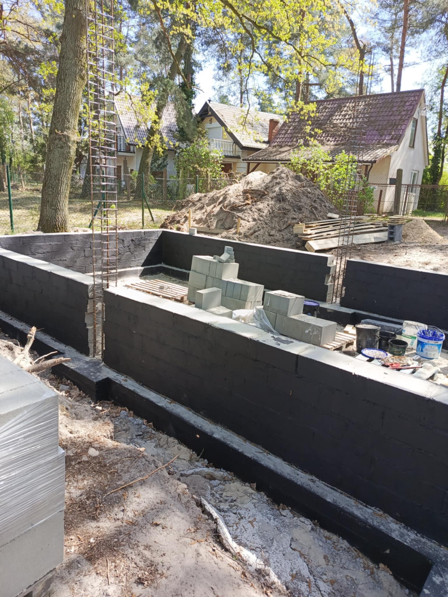 Fundamenty domu w trakcie budowy: czarne ściany fundamentowe, wystające pręty zbrojeniowe, paleta z bloczkami betonowymi, narzędzia, domy w tle, słoneczny dzień.