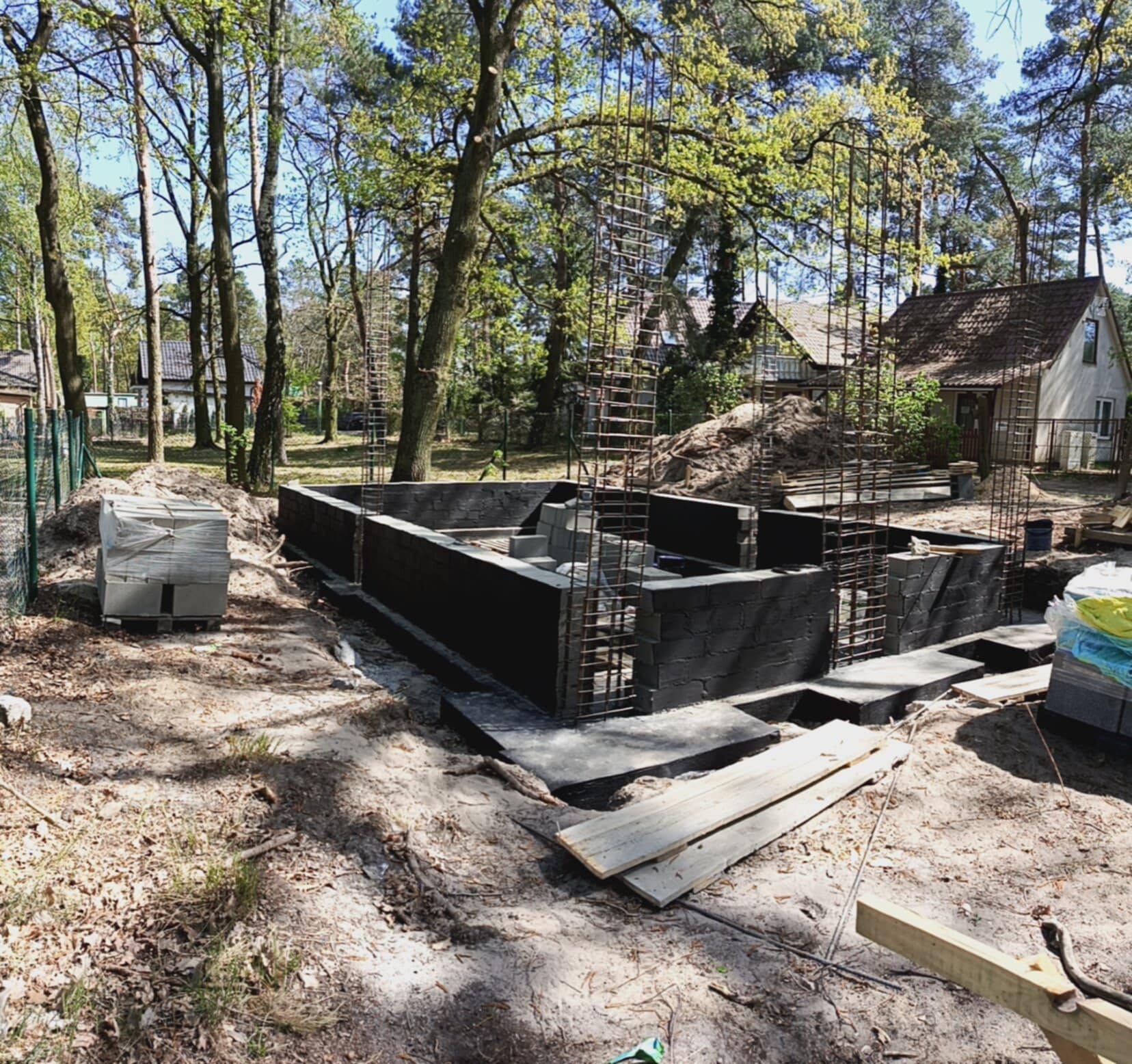 Fundamenty z bloczków betonowych i zbrojeniami, otoczone drzewami. W tle widoczny dom jednorodzinny. Etap budowy w naturalnym otoczeniu.