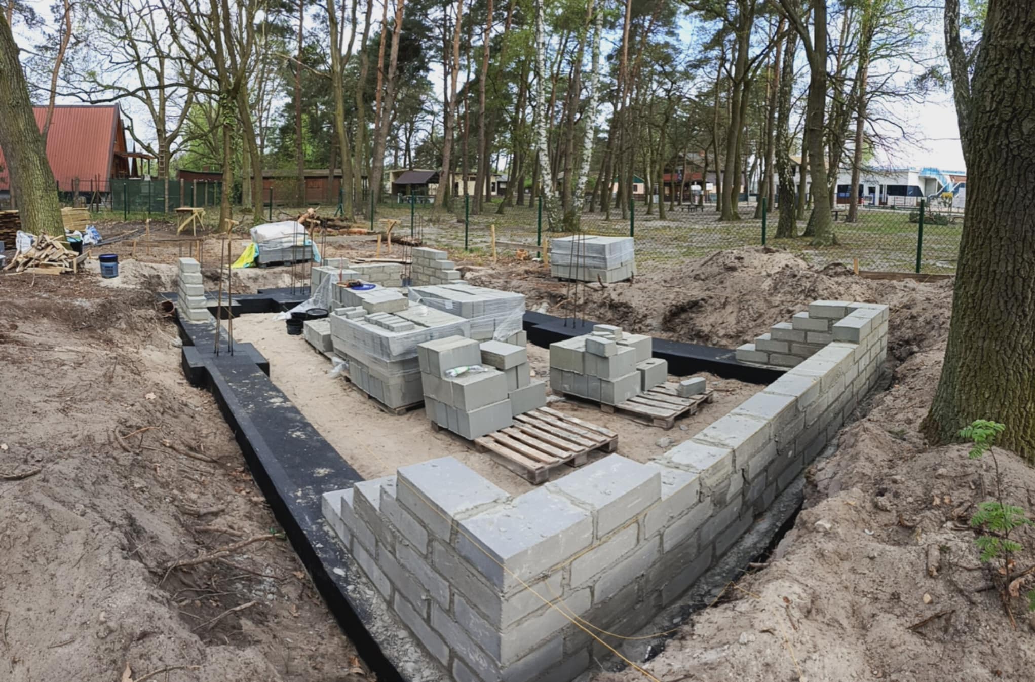 Wykop pod fundamenty domu z częściowo wymurowanymi ścianami z bloczków betonowych. Widoczne izolacje i zapasy materiałów budowlanych na placu budowy w otoczeniu drzew.