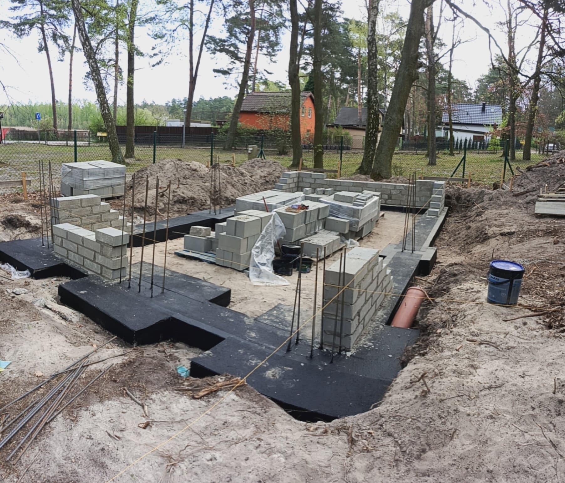 Budowa fundamentów domu: izolacja czarną folią, częściowo wymurowane ściany z szarego bloczka betonowego, wystające pręty zbrojeniowe, otoczenie ziemne.