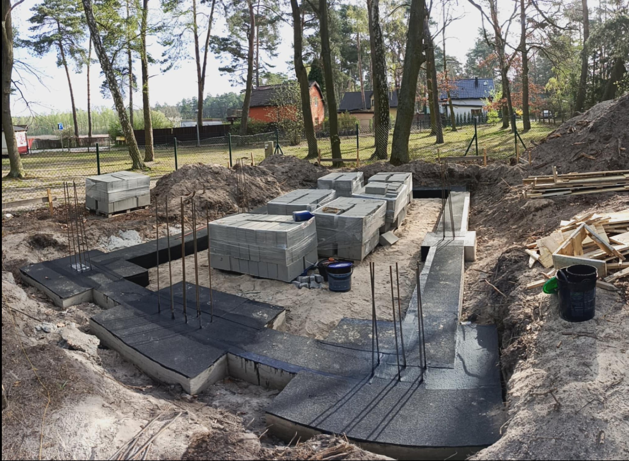 Zalane fundamenty z wystającymi prętami zbrojeniowymi, zabezpieczone czarną izolacją, na tle drzew i zabudowań. Na ziemi ułożone bloczki betonowe i narzędzia.