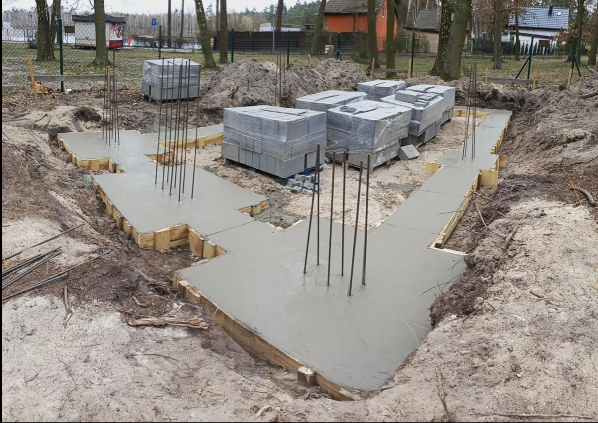 Zalane fundamenty domu z wystającymi prętami zbrojeniowymi i bloczkami betonowymi w folii. Widok z góry na wykopane fundamenty w ziemi.