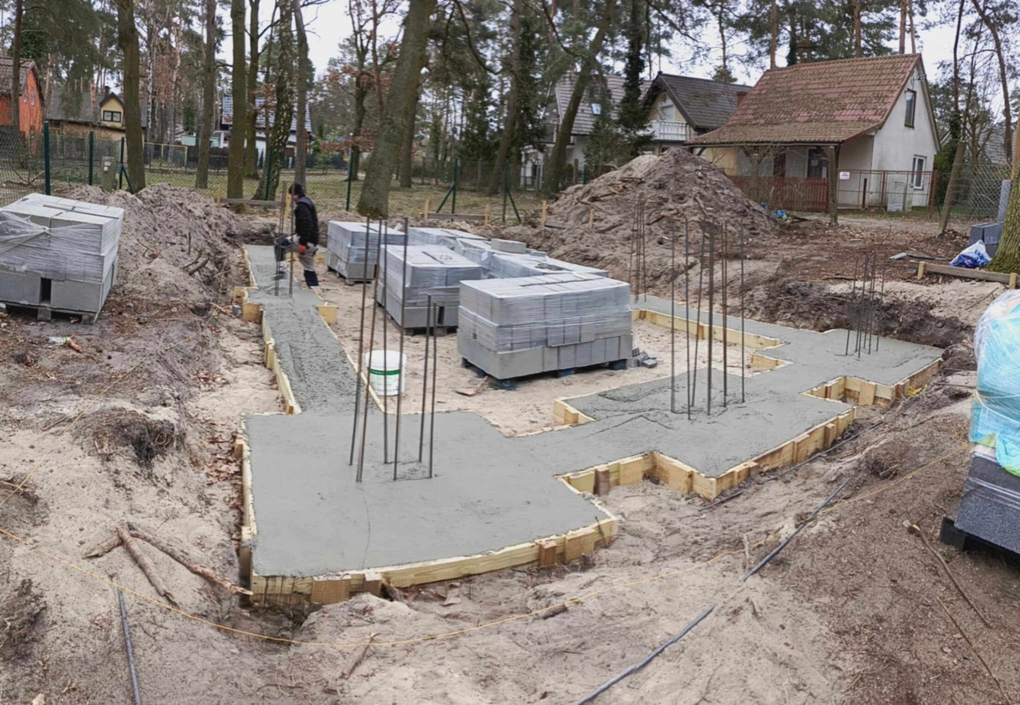 Wylane fundamenty pod budowę domu, widoczne szalunki, zbrojenie i bloczki betonowe na paletach. W tle domy i drzewa.