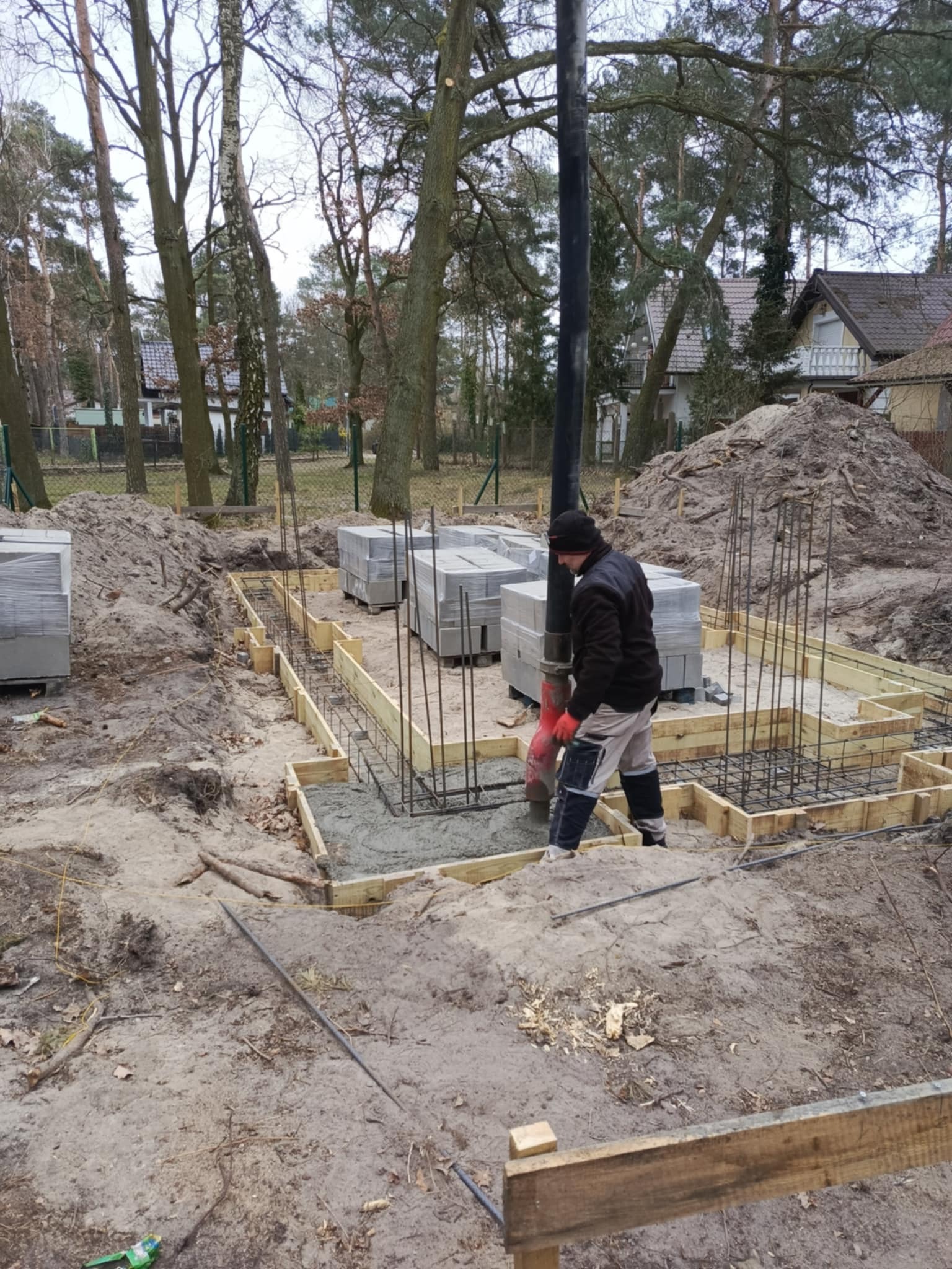 Fundamenty domu w Lubiczu: wylewanie betonu z pompy do szalunków zbrojeniowych. Na placu budowy widoczne bloczki i ziemia.