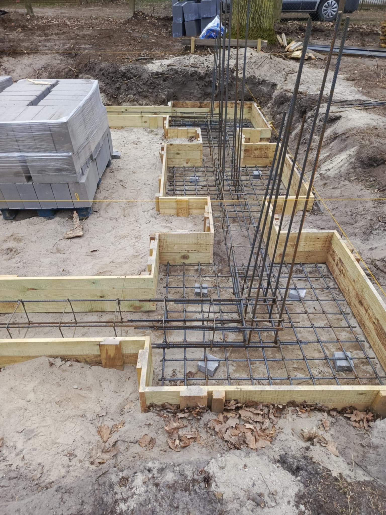 Szczegółowy widok na szalunek fundamentów zbrojonych stalą, przygotowany do wylania betonu. Na pierwszym planie paleta bloczków fundamentowych.