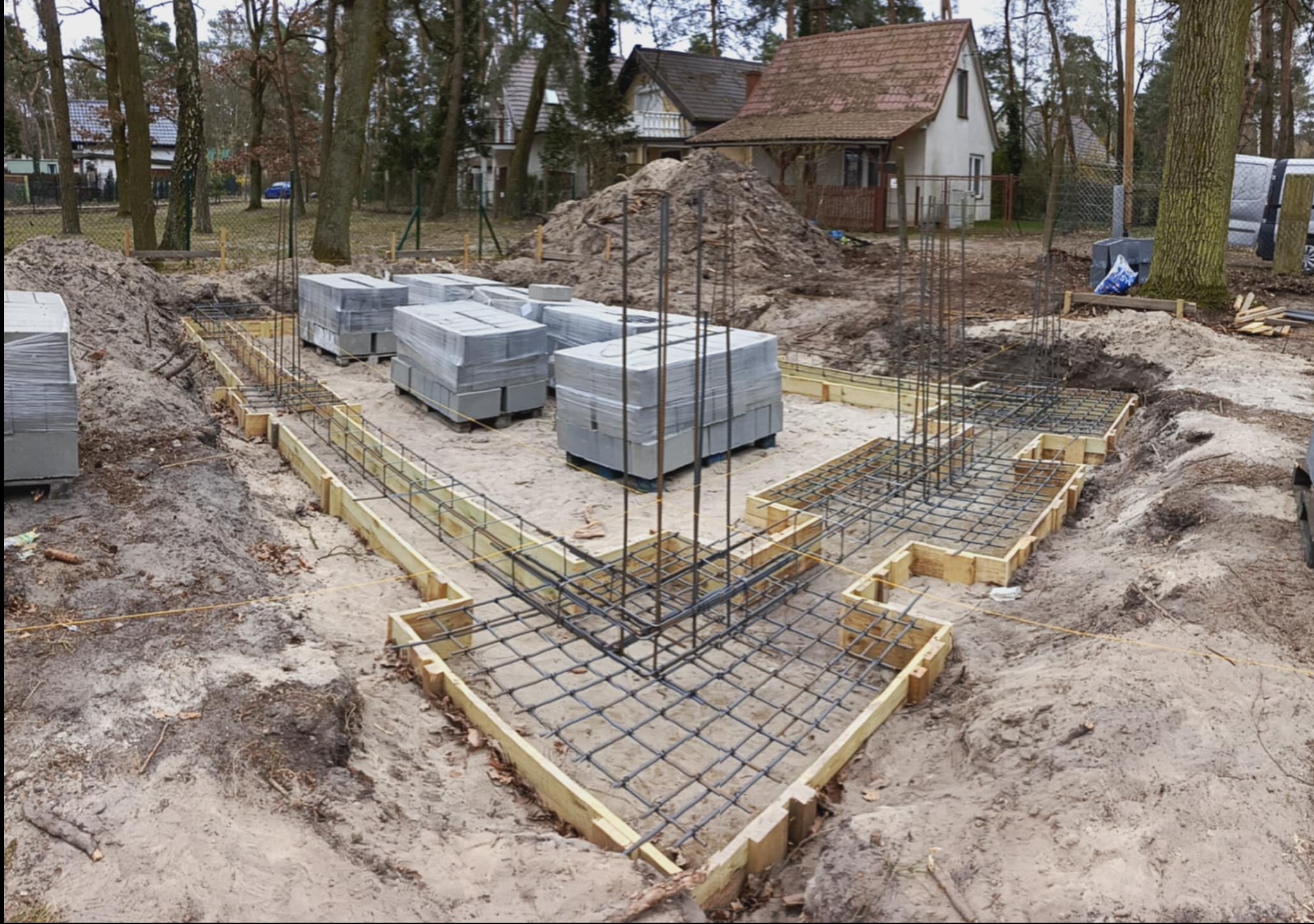 Przygotowanie fundamentów pod budowę domu: szalunki zbrojeniowe i bloczki fundamentowe na działce z drzewami w tle, gotowe do zalania betonem.