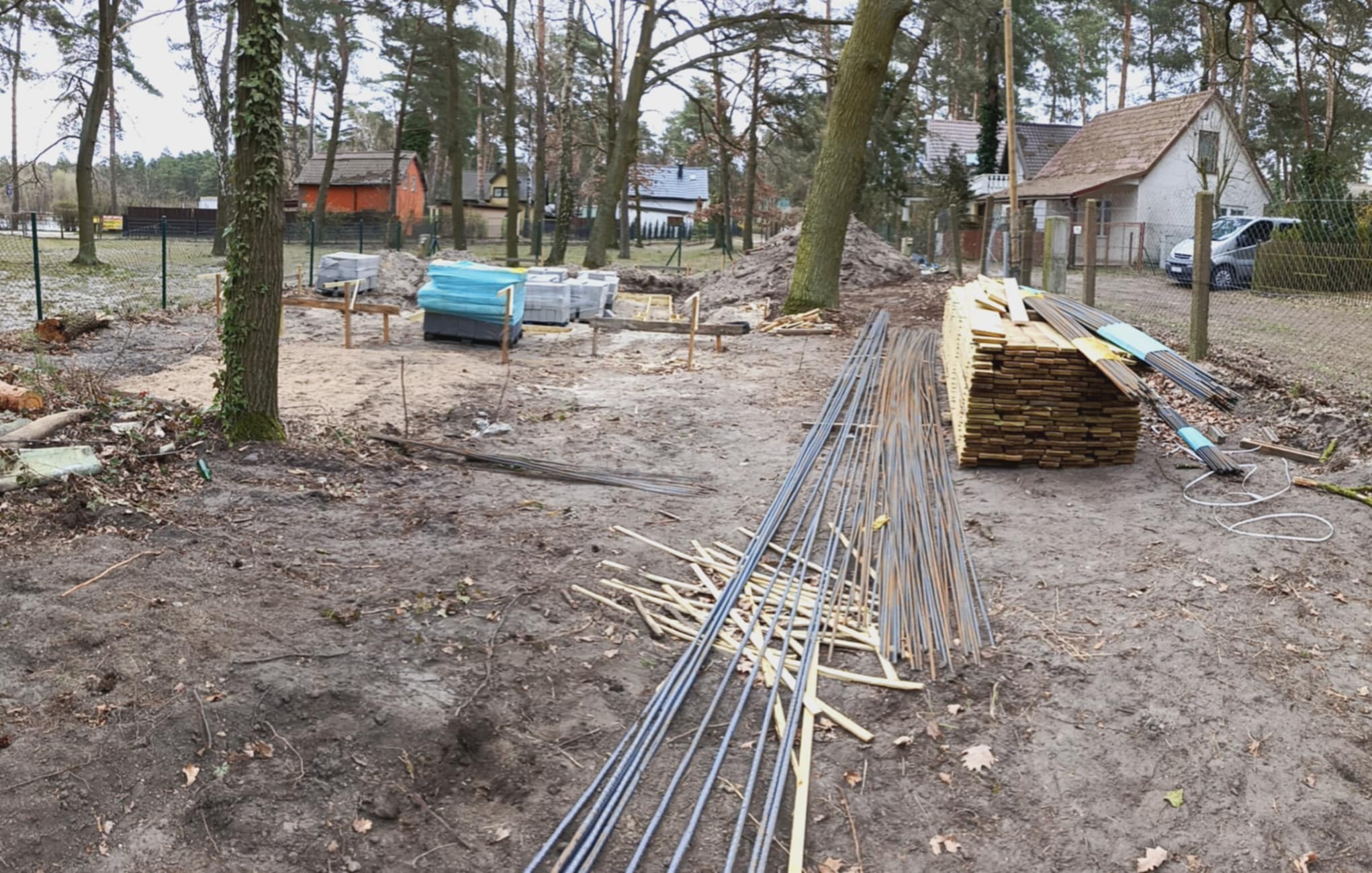 Plac budowy w lesie: zbrojenie, drewno, bloczki betonowe. Przygotowania do wylania fundamentów pod budynek mieszkalny. Widoczne domy w tle.