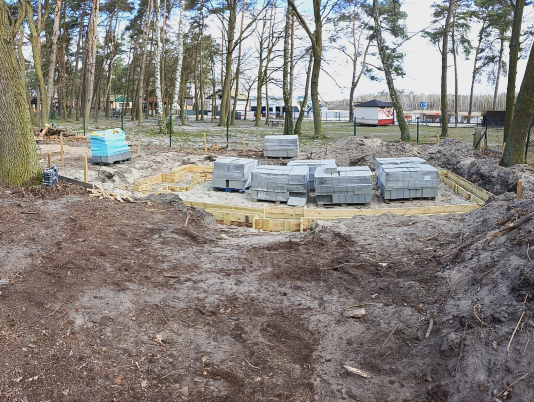 Fundamenty pod budowę domu w lesie: szalunek z desek, bloczki betonowe na paletach, teren przygotowany pod wylewkę. Widoczne drzewa i budynki w tle.
