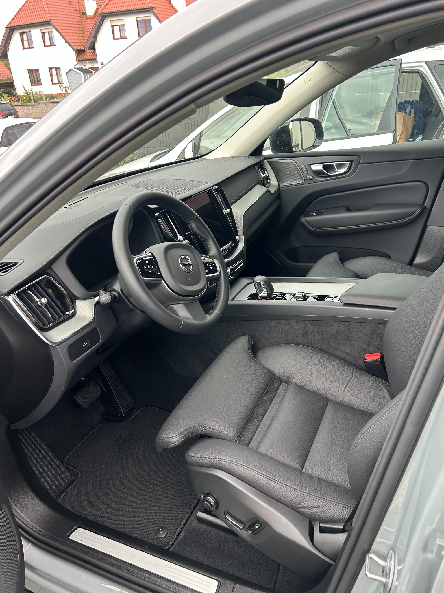 Wnętrze szarego Volvo XC60 z czarną skórzaną tapicerką, widok z zewnątrz przez otwarte drzwi, zarys budynków w tle. Czyste i zadbane wnętrze.