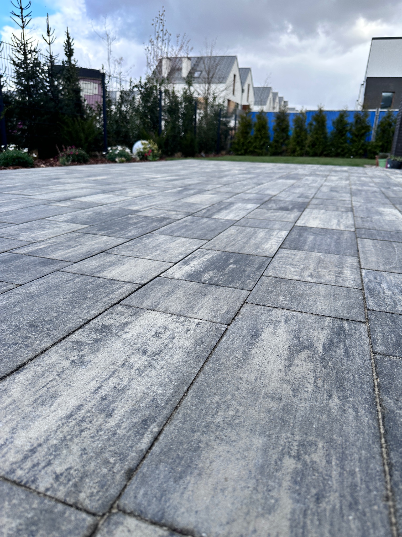 Szara kostka brukowa ułożona na posesji z widokiem na domy i zieleń. Widoczne geometryczne wzory i faktura kostki. Całość tworzy uporządkowaną przestrzeń.