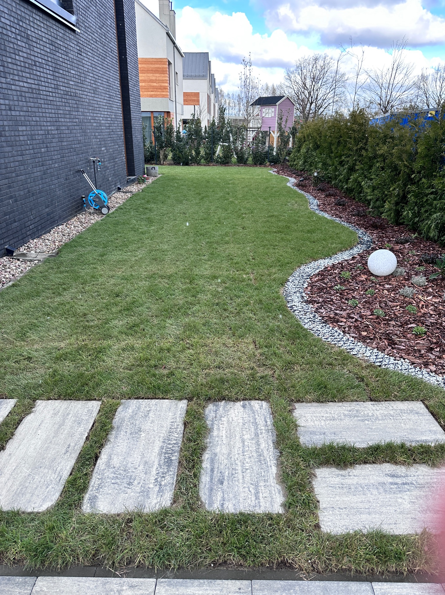Uporządkowany ogród z zielonym trawnikiem, ścieżką z płyt i dekoracyjną rabatą z kulą. W tle budynki mieszkalne i niebieski wąż ogrodowy na ścianie domu.