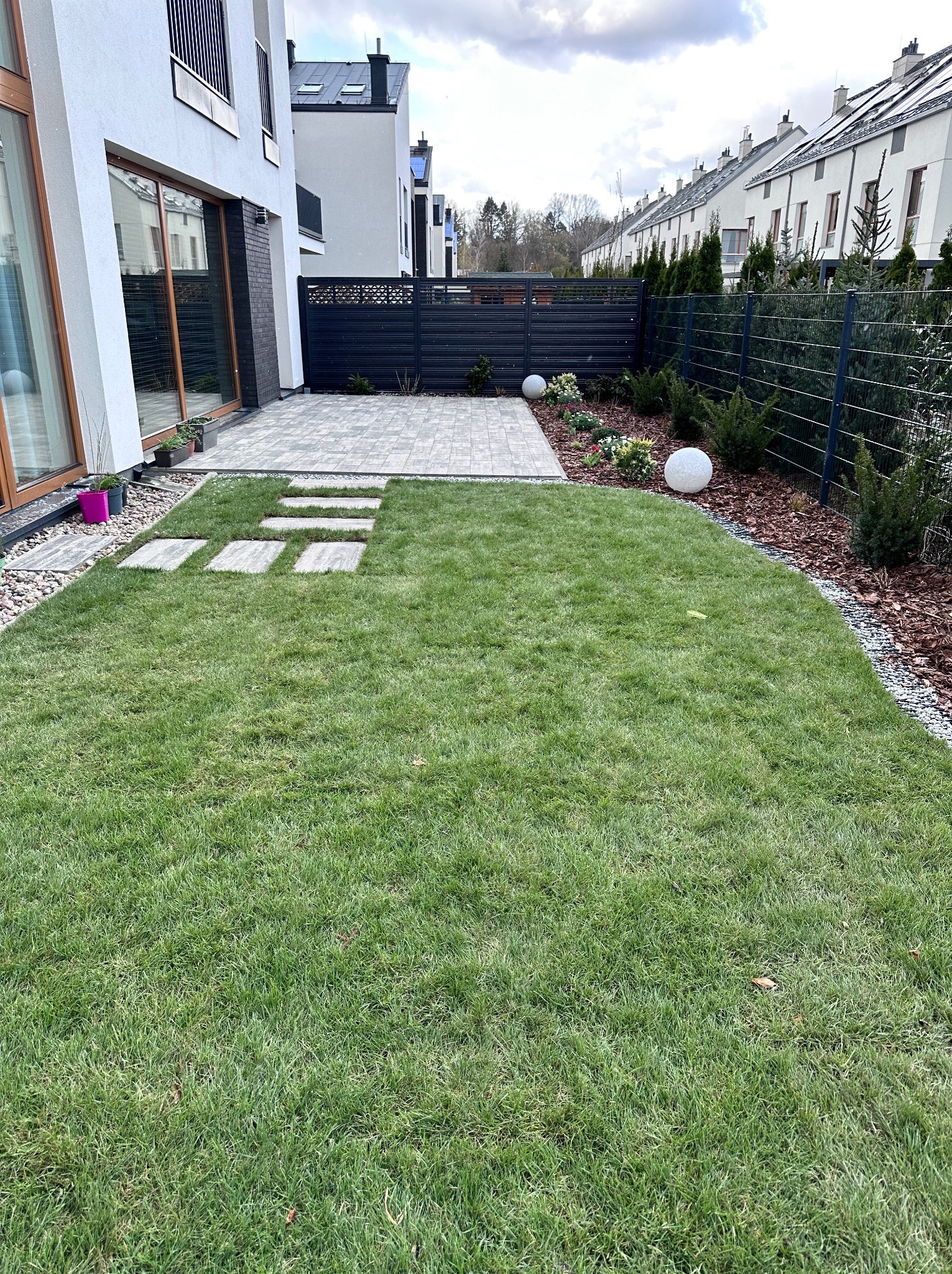 Nowoczesny ogród z trawnikiem, ścieżką z płyt i kostki brukowej. Widoczne elementy dekoracyjne, takie jak kule ogrodowe i rabaty z roślinami. Projekt zagospodarowania przestrzeni.