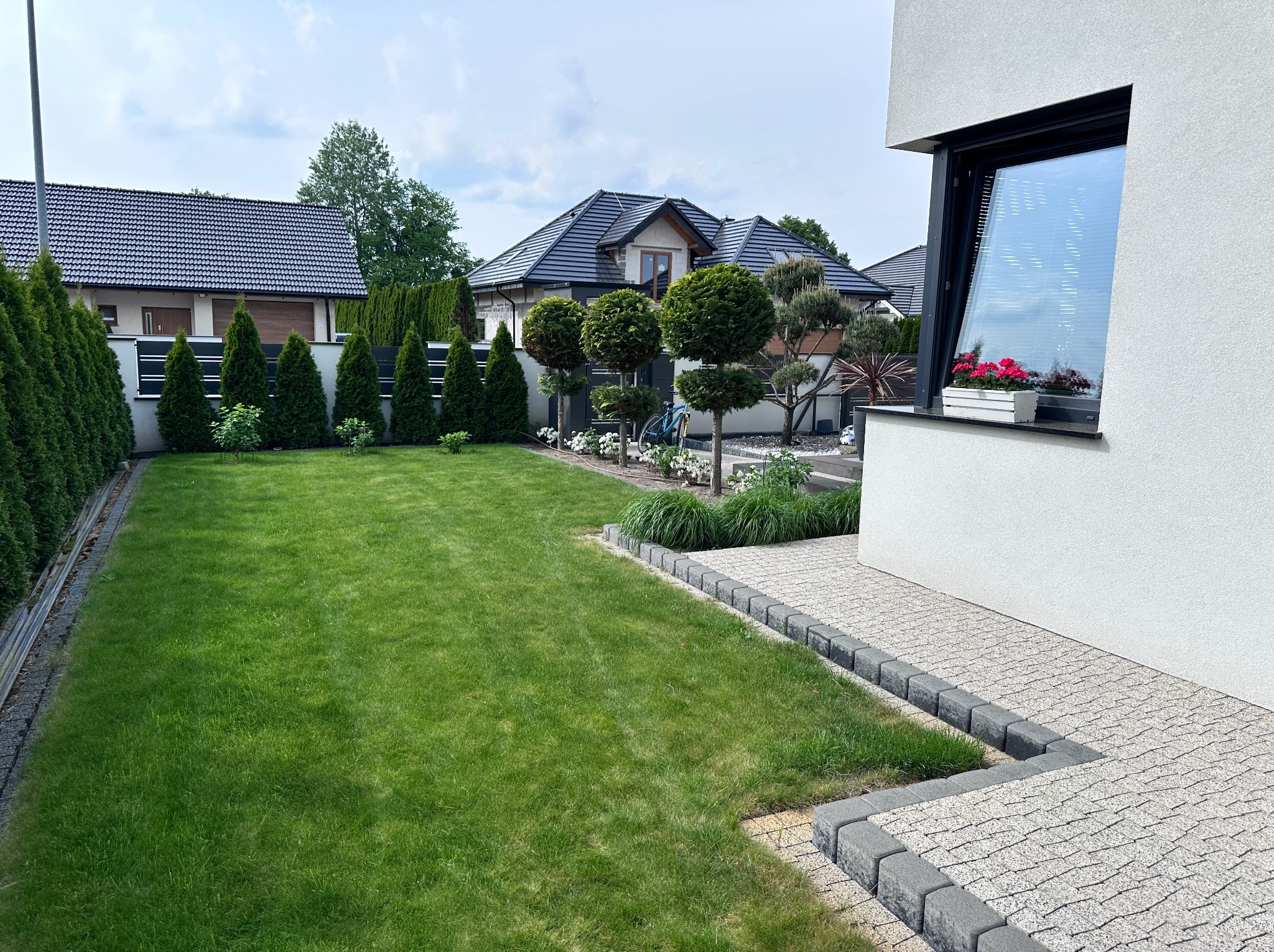 Zadbany ogród z równo przystrzyżonym trawnikiem, ozdobnymi drzewkami i kostką brukową wokół domu. Widok na domy w tle i błękitne niebo.