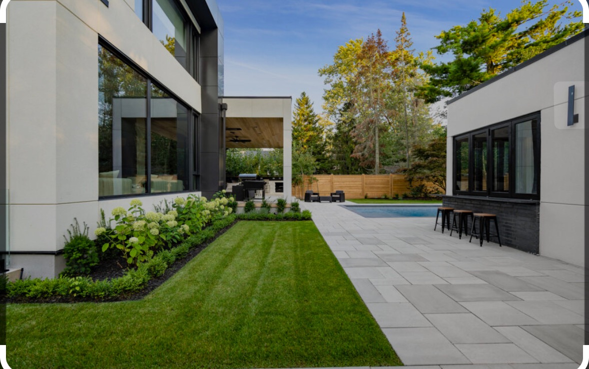 Nowoczesny ogród z równo przystrzyżonym trawnikiem, kostką brukową i minimalistyczną architekturą. Widok na basen i zielone drzewa w tle.