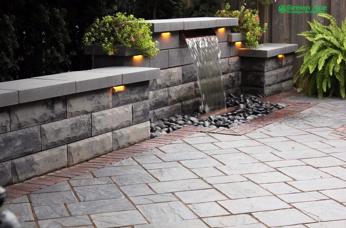 Ogród z kaskadą wodną, podświetlonymi roślinami i brukowaną ścieżką. Kamienna ściana z wbudowanym oświetleniem. Elementy z kostki brukowej w różnych odcieniach szarości i czerwieni.