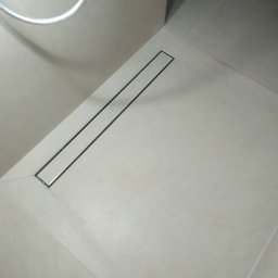 RAKAS - Minimalistyczna łazienka z odpływem liniowym w posadzce, wykończona jasnymi płytkami. Ujęcie z góry, widoczny fragment baterii prysznicowej.