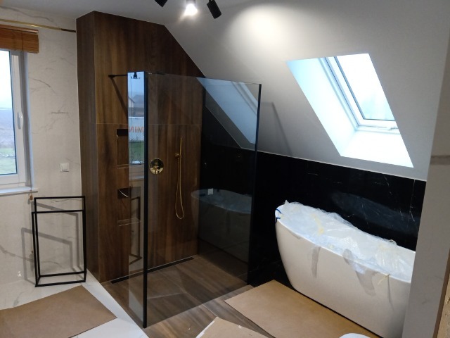 Nowoczesna łazienka na poddaszu ze szklaną kabiną prysznicową, wolnostojącą wanną pod oknem dachowym i marmurowymi płytkami. Widoczne detale wykończeniowe.