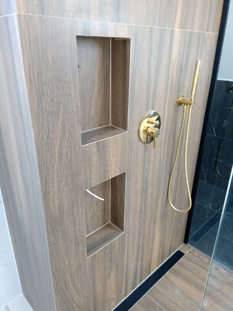 Nowoczesna kabina prysznicowa z wnękami wyłożonymi płytkami imitującymi drewno, złota armatura, czarny odpływ liniowy i szklana ściana kabiny.