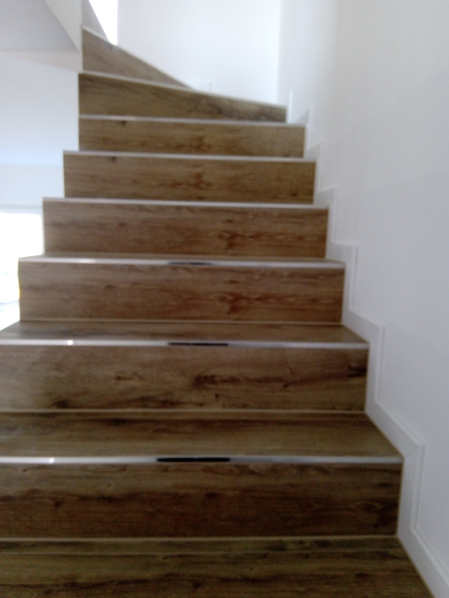Wykończone schody z drewnopodobnymi płytkami i srebrnymi listwami antypoślizgowymi, prowadzące w górę w jasnym wnętrzu.